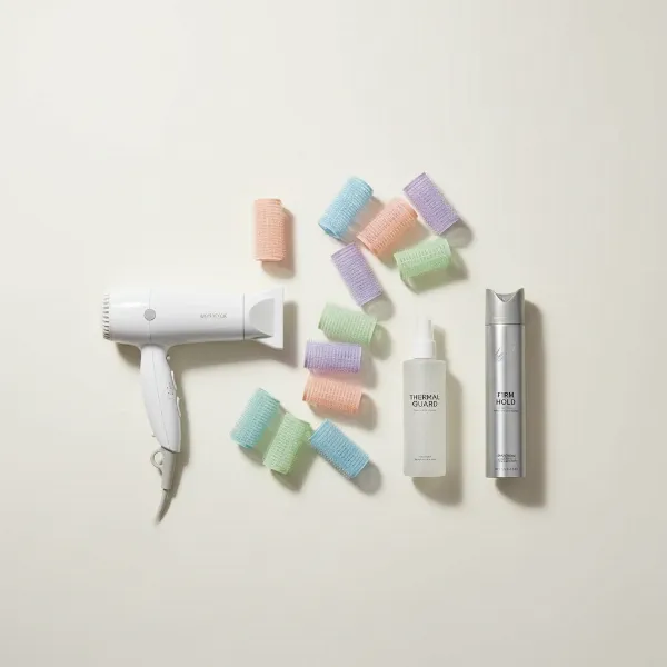 A flat lay image showing various sizes of velcro rollers, a hair dryer with a concentrator nozzle, and styling products like heat protectant and hairspray laid out on a clean surface for a hair styling tutorial.